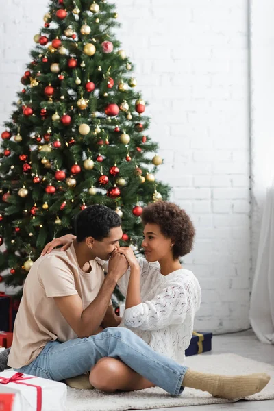 Молодой африканский американец целует руку сексуальной женщины, сидящей на полу возле рождественской елки — стоковое фото