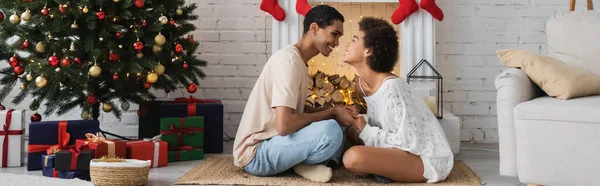 Vista lateral de la feliz pareja afroamericana cogida de la mano en el suelo cerca del árbol de Navidad y la chimenea, pancarta - foto de stock