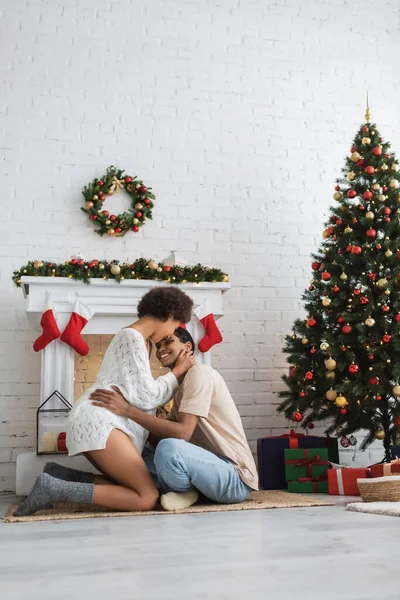 Jovem casal afro-americano abraçando perto da árvore de natal e lareira decorada na sala de estar — Fotografia de Stock