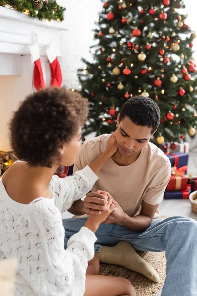 Jeune homme afro-américain tenant la main d'une petite amie touchant son cou près de l'arbre de Noël flou — Photo de stock