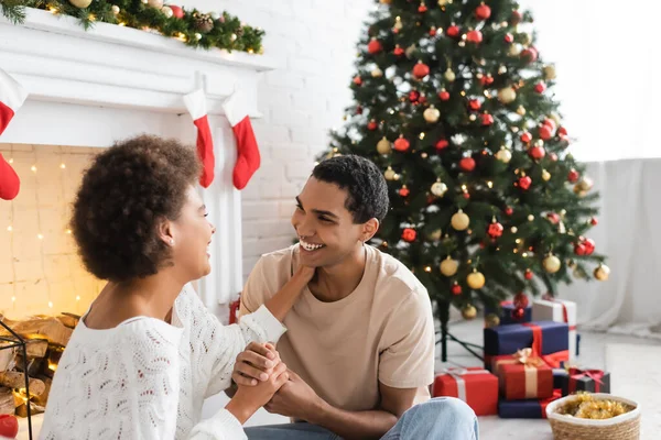 Alegre pareja afroamericana tomados de la mano y mirándose el uno al otro cerca de árbol de Navidad y chimenea - foto de stock