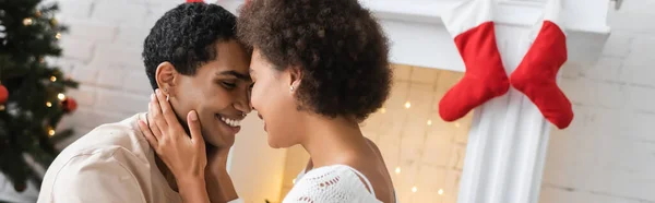 Jeune femme afro-américaine embrassant le visage du petit ami tout en célébrant Noël à la maison, bannière — Photo de stock