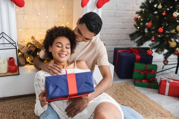 Heureuse femme afro-américaine avec boîte cadeau assis près du jeune petit ami et cheminée à la maison — Photo de stock