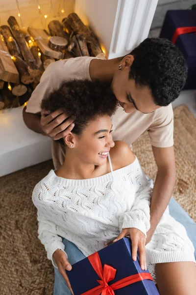 Giovane uomo africano americano toccare i capelli ricci della fidanzata felice seduto con scatola regalo vicino al camino — Foto stock