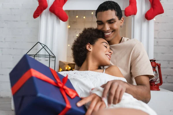 Сексуальная женщина с подарком на Рождество рядом с улыбающимся африканским американским парнем, сидящим у камина — стоковое фото