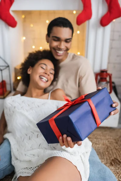 Alegre mujer afroamericana sosteniendo caja de regalo azul con cinta roja cerca de novio sobre fondo borroso - foto de stock