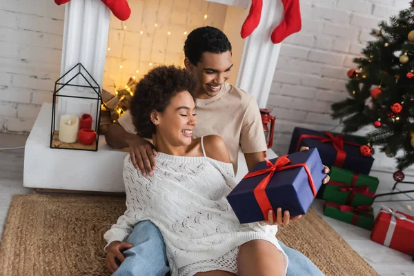 Alegre casal afro-americano com presente de Natal sentado no chão perto da lareira — Fotografia de Stock