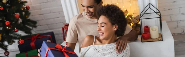 Smiling african american couple looking at christmas present while sitting near fireplace at home, banner — Stock Photo