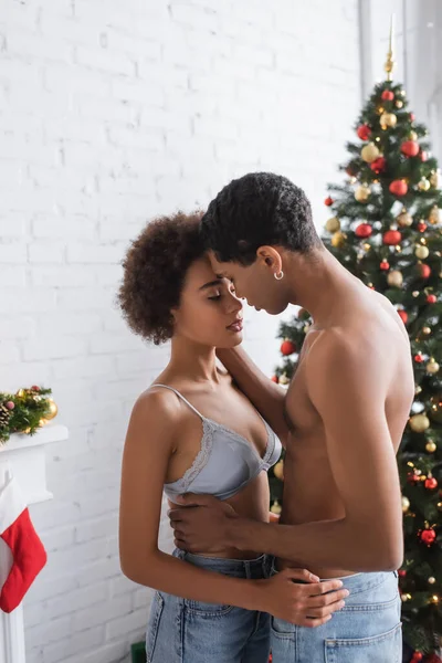 Sensual afroamericana mujer en sujetador y sin camisa novio abrazándola cerca de árbol de Navidad - foto de stock