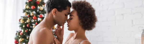 Side view of curly african american woman touching lips of sexy boyfriend near blurred christmas tree, banner — Stock Photo