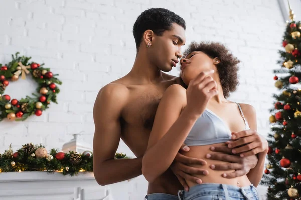 Jovem e sexy afro-americano homem abraçando mulher sedutora perto de enevoado Natal decoração — Fotografia de Stock