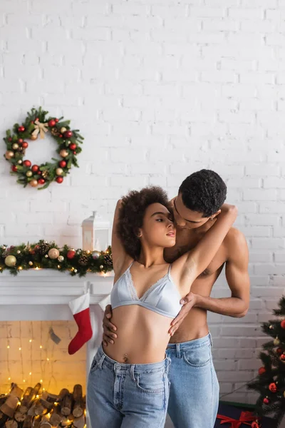 Young african american man hugging girlfriend in bra and jeans near fireplace with christmas decoration — Stock Photo
