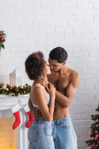 Homem americano Africano sem camisa em jeans abraçando namorada apaixonada perto de lareira decorada — Fotografia de Stock