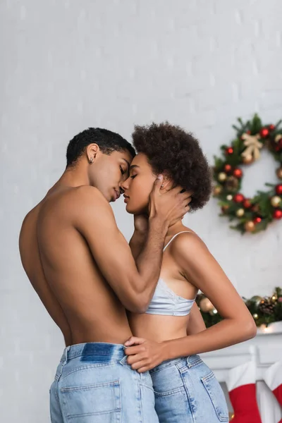 Shirtless muscular man kissing seductive african american girlfriend near blurred christmas wreath — Stock Photo