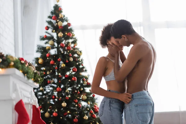 Árbol de navidad decorado cerca de pareja afroamericana sexy besándose en casa - foto de stock