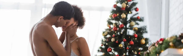 Без сорочки афроамериканський чоловік цілує сексуальну молоду жінку біля ялинки, банер — стокове фото