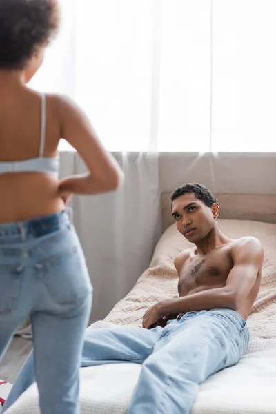 Африканский мужчина без рубашки лежит на кровати в джинсах и смотрит на сексуальную девушку на размытом переднем плане — стоковое фото