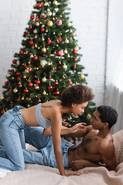 Вид на сексуальную африканскую женщину в джинсах и лифчике, соблазняющую мужчину без рубашки на кровати возле рождественской елки — стоковое фото
