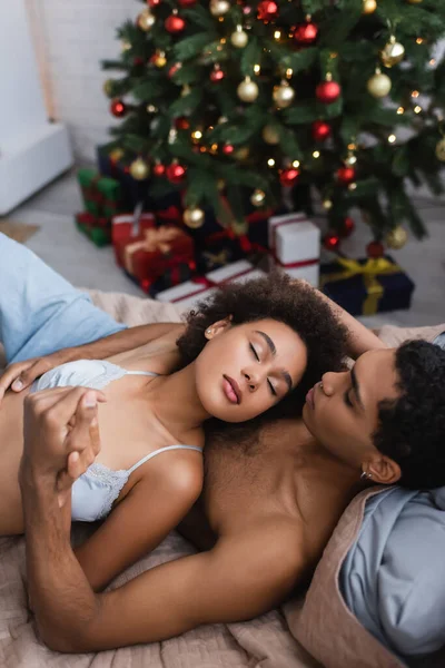 Visão de alto ângulo de casal americano africano sexy de mãos dadas na cama perto de árvore de natal borrada — Fotografia de Stock