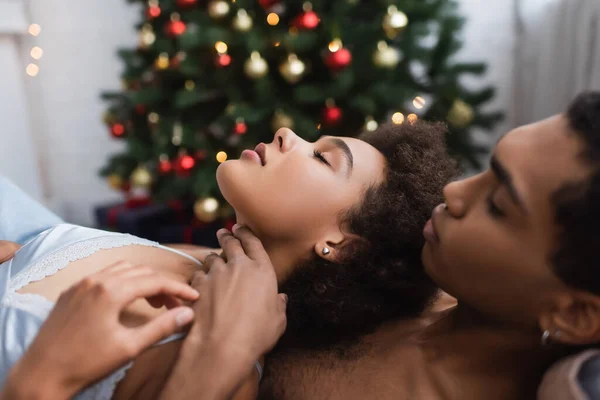 Vista lateral da mulher americana africana sensual no sutiã perto do jovem namorado e árvore de natal borrada — Fotografia de Stock