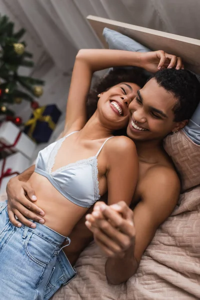 Vista superior do casal americano africano sexy de mãos dadas e rindo enquanto deitado na cama — Fotografia de Stock