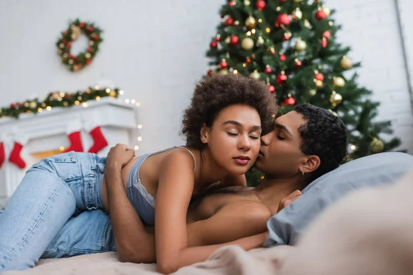 Appassionata donna afroamericana ad occhi chiusi che abbraccia l'uomo in camera da letto con decorazioni natalizie sfocate — Foto stock