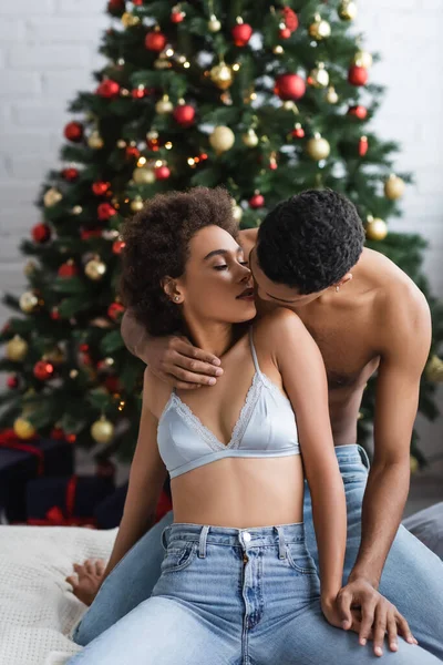 Africano americano hombre besos sexy mujer en sujetador y jeans en cama cerca de navidad árbol - foto de stock