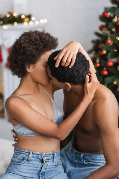 Junges und leidenschaftliches afrikanisch-amerikanisches Paar in Jeans küsst sich in der Nähe von verschwommenem Weihnachtsdekor — Stockfoto