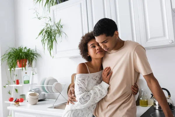 Sexy africana americana mujer en blanco punto suéter sonriendo cerca joven novio en cocina - foto de stock