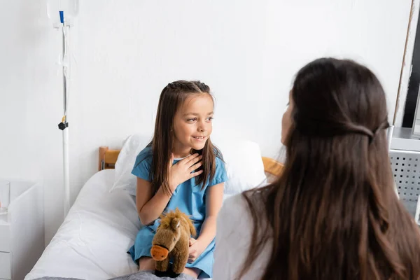 Smiling girl holding soft toy and touching chest near doctor in hospital ward — Stock Photo