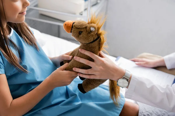 Vista recortada del pediatra sosteniendo juguete suave cerca del niño en la cama del hospital - foto de stock
