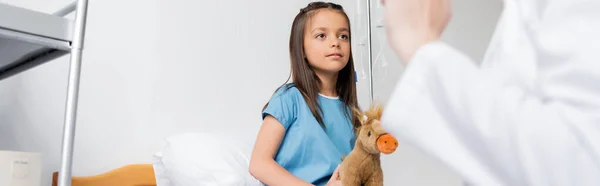 Niño con juguete suave mirando al pediatra borroso en la sala del hospital, pancarta - foto de stock