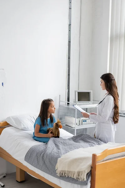 Vista laterale del pediatra guardando appunti vicino alla ragazza sul letto d'ospedale — Foto stock
