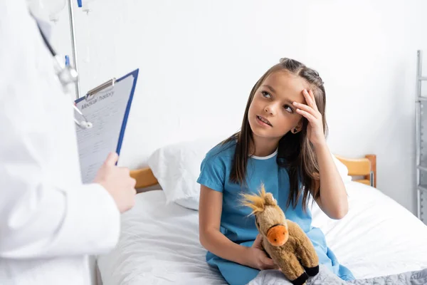Girl holding soft toy and touching head near blurred pediatrician with clipboard in clinic — Stock Photo