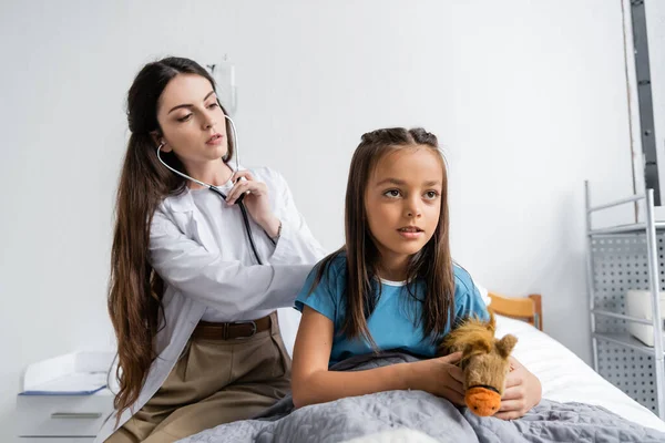 Ребенок с мягкой игрушкой сидит рядом с доктором со стетоскопом на больничной койке — стоковое фото