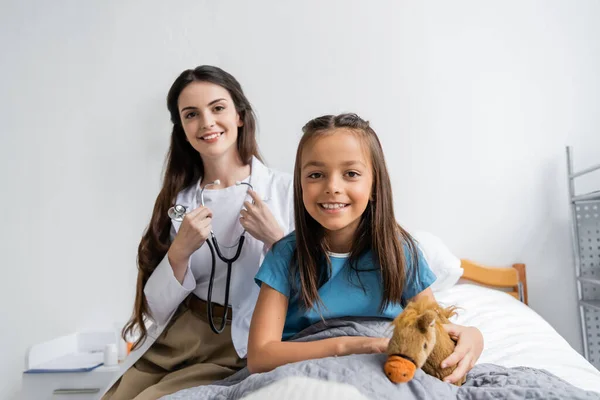 Smiling kid holding soft toy near doctor with stethoscope looking at camera in hospital ward — Stock Photo