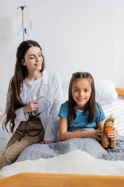 Médecin souriant tenant un stéthoscope près d'un enfant avec un jouet sur un lit d'hôpital — Photo de stock