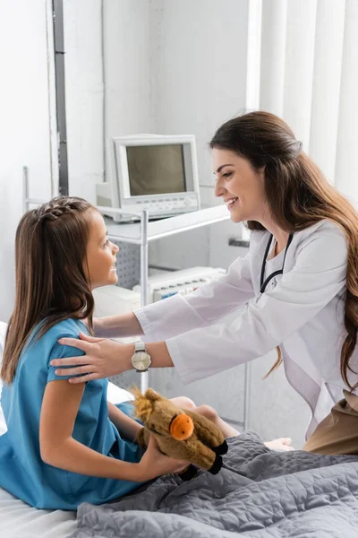 Вид сбоку улыбающийся доктор смотрит на ребенка с игрушкой на больничной койке — стоковое фото