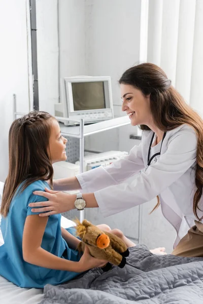 Side view of smiling doctor looking at kid with toy on hospital bed — Stock Photo