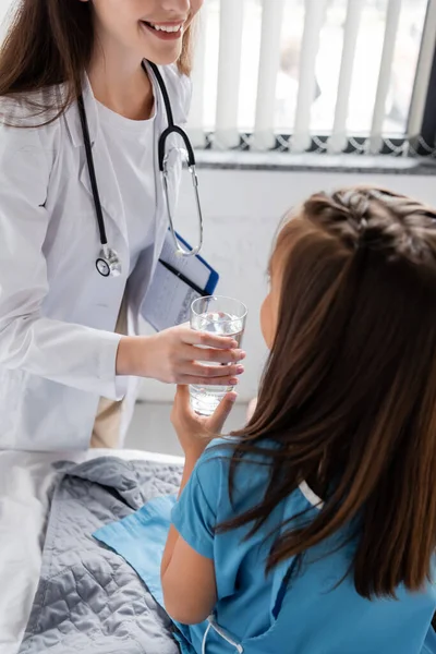 Pediatra sorrindo dando vidro de água para menina em vestido de paciente na cama clínica — Fotografia de Stock