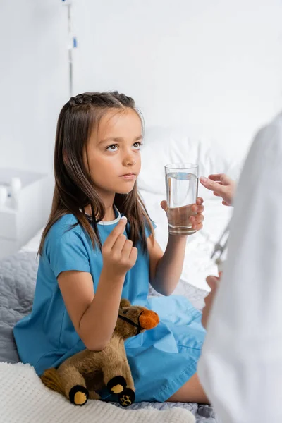Pediatra borroso de pie cerca de un niño con vaso de agua y píldora en la cama en la clínica - foto de stock