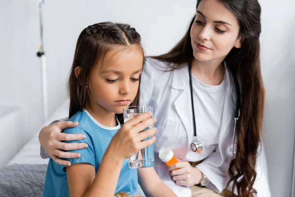 Pédiatre tenant des pilules près de l'enfant avec un verre d'eau dans la salle d'hôpital — Photo de stock