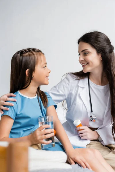 Médico sonriente abrazando a chica con vaso de agua y sosteniendo pastillas en la sala de hospital - foto de stock