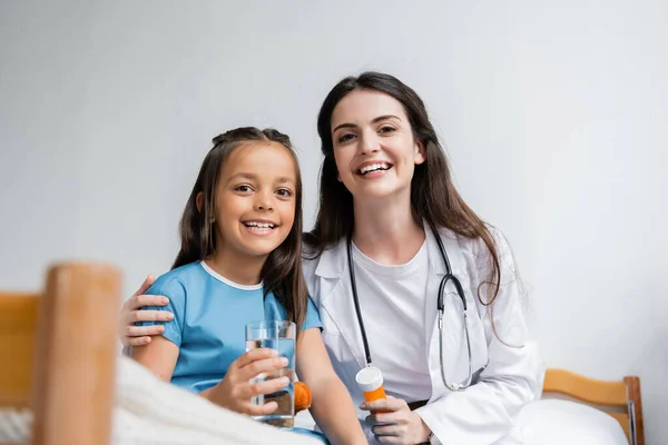 Pédiatre souriant et enfant en robe de patient tenant un verre d'eau et des pilules dans la salle d'hôpital — Photo de stock