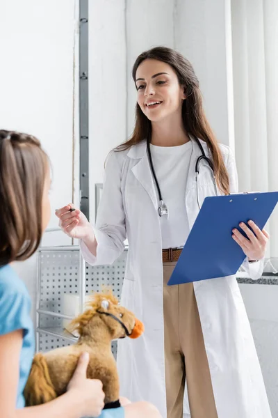 Усміхнений лікар тримає кишеню і розмовляє з дитиною з іграшкою в лікарняному відділенні — стокове фото