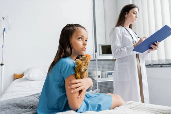 Girl in patient gown holding toy while sitting on hospital bed near doctor — Stock Photo