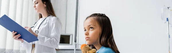 Niño con juguete mirando hacia otro lado cerca borroso pediatra sosteniendo portapapeles en la clínica, pancarta - foto de stock
