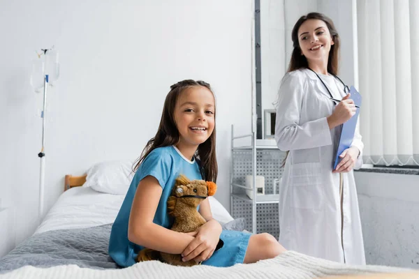 Sonriente chica con juguete suave mirando a la cámara cerca borrosa médico en la sala de hospital - foto de stock