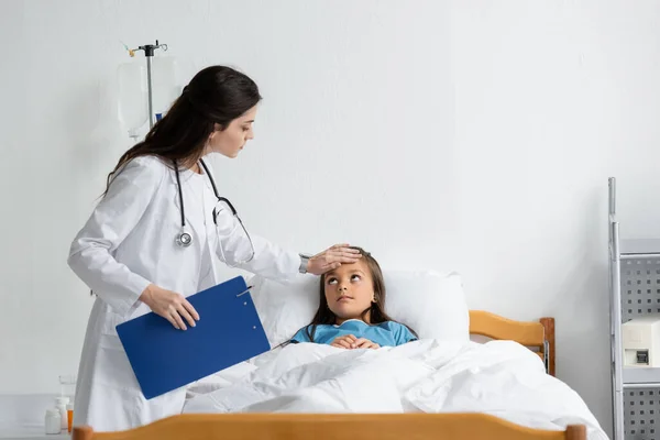 Doctor with clipboard examining forehead of child in patient gown on hospital bed — Stock Photo