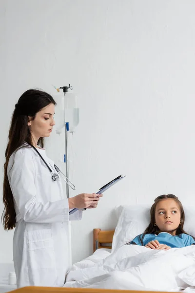 Docteur regardant presse-papiers près de l'enfant en robe de patient sur le lit à l'hôpital — Photo de stock